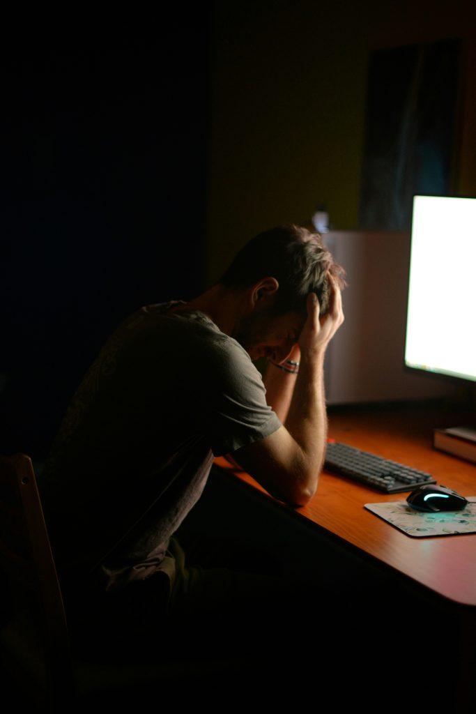 Man despairing: sitting in front of a bright computer screen, head in hands in a dark room.